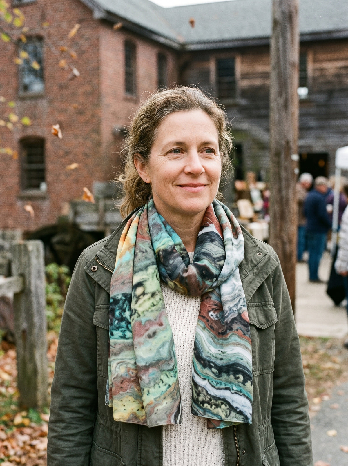 A woman wearing a dark olive green jacket and a colorful marbled scarf with green, blue, orange, and black patterns, standing outdoors in front of a brick building.