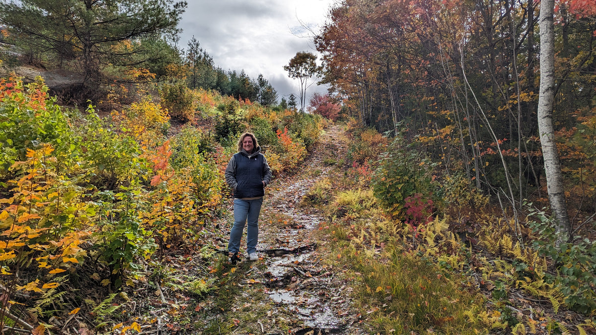 A person standing on a muddy forest path surrounded by colorful autumn foliage in shades of yellow, orange, and red under an overcast sky.